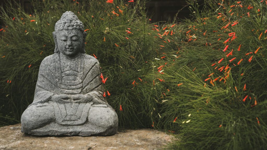 Stone buddha sits in front of tall green grass with orange flowers