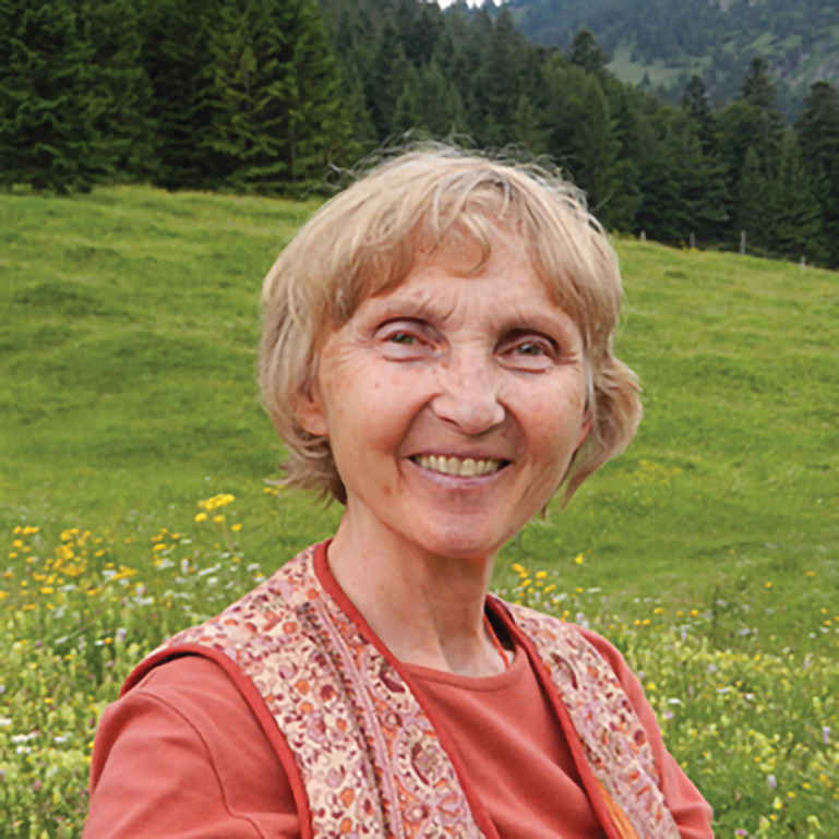 A woman smiles while wearing a pink shirt with a pink patterned vest on top. There is a field in the background with many trees. 