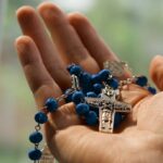 A hand holds a blue Catholic rosary.