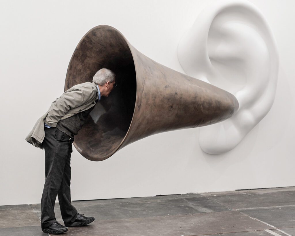 A man is depicted leaning his head into a large sculpture of an ear and ear-horn