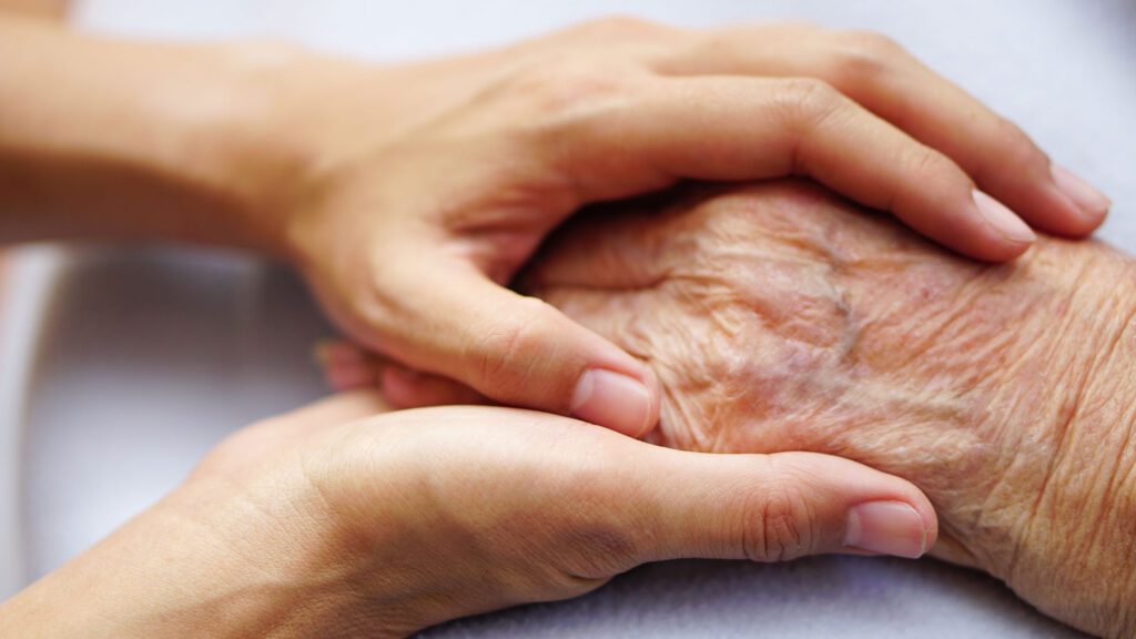 A family member holding a elderly woman’s hand by hospital bed.