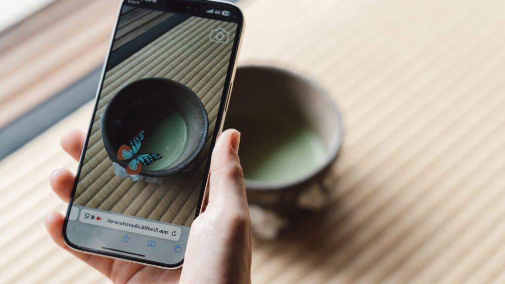 A hand holds a smartphone displaying a photo of a matcha bowl with a digital butterfly overlay. The real bowl, filled with green tea, sits on a tatami mat.